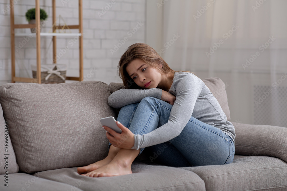 Portrait of depressed young woman sitting on couch with smartphone in her hands Portrait of depressed young woman sitting on couch with smartphone in her hands