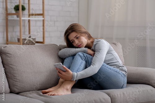 Portrait of depressed young woman sitting on couch with smartphone in her hands