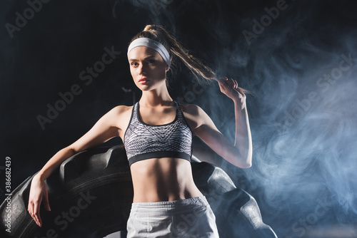 Selective focus of sportswoman touching hair near tire in sports center with smoke