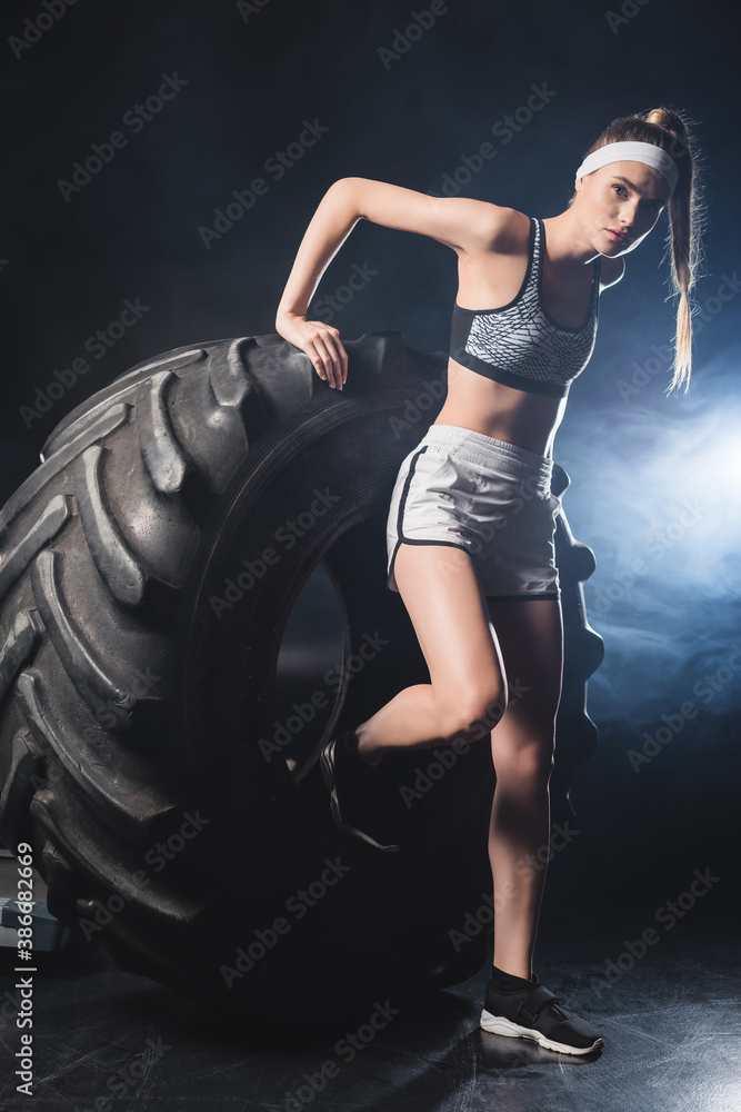 Sportswoman standing near tire in gym with smoke