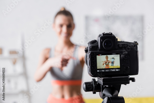 Selective focus of smiling sportswoman pointing at sports bottle near digital camera