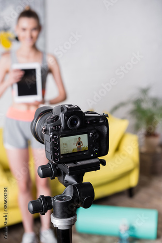 Selective focus of smiling sportswoman with digital tablet standing near digital camera at home