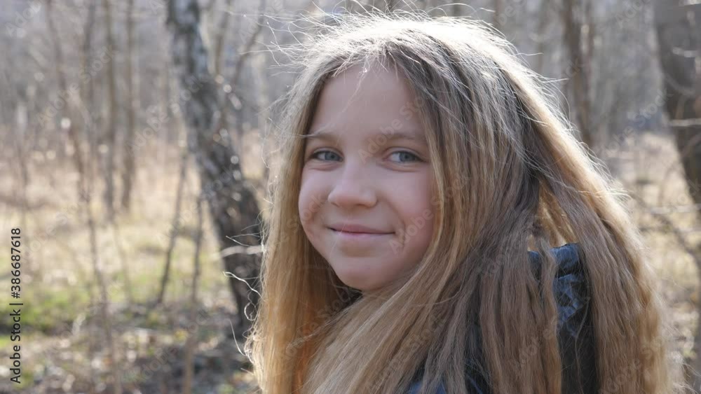 Portrait of happy small child against the blurred background of a forest in early spring. Smiling blonde girl looking into camera outdoor. Close up emotions of female kid with glad expression on face Portrait of happy small child against the blurred background of a forest in early spring. Smiling blonde girl looking into camera outdoor. Close up emotions of female kid with glad expression on face