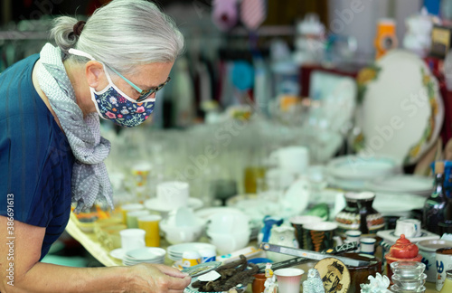 A senior woman with glasses and scarf wearing a medical mask due to coronavirus while looking at home accessories at the flea market.