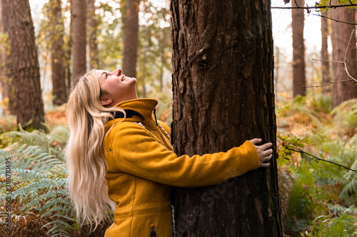 Young blonde woman in yellow coat hugging a tree in the forest and looking up to the tree top