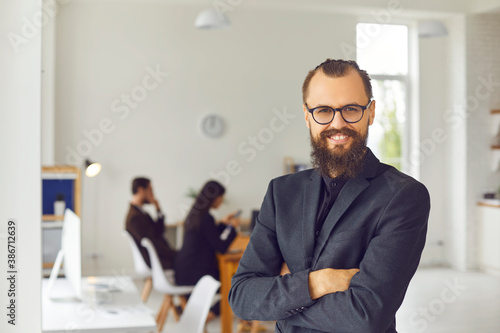 Happy charismatic young business leader standing arms crossed against blurred office background