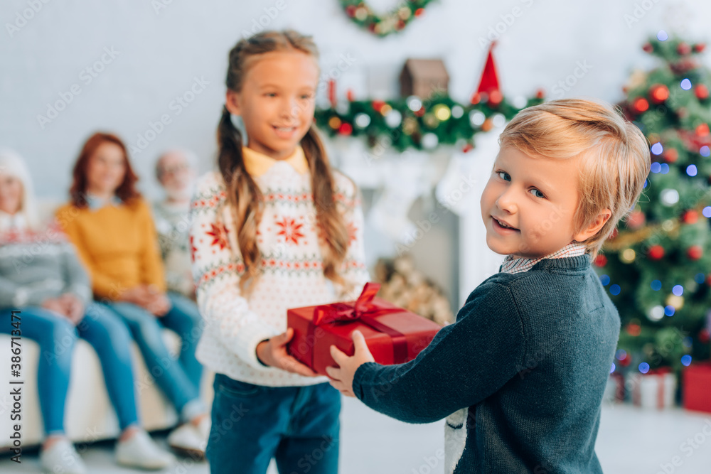 Obraz premium smiling boy looking at camera while presenting christmas gift to happy sister