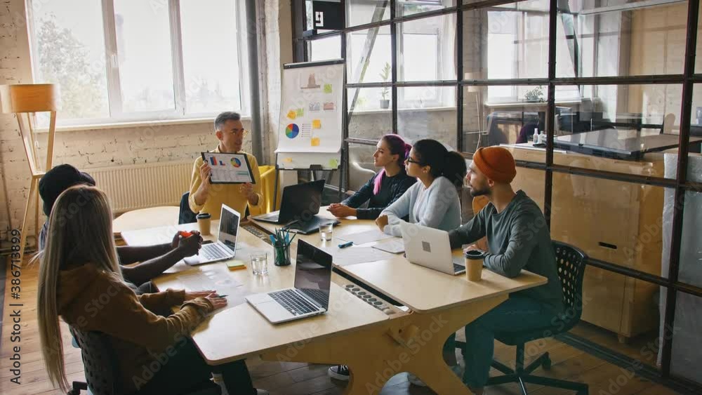 Mature team leader making presentation for colleagues in office, showing colorful chart, sitting in office Mature team leader making presentation for colleagues in office, showing colorful chart, sitting in office