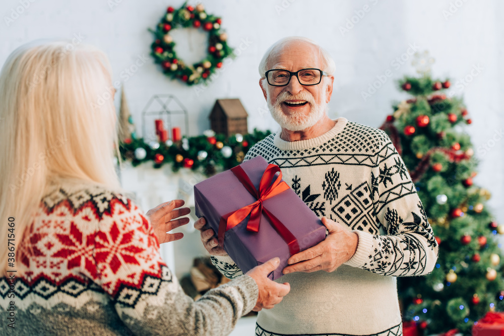 selective focus of happy senior man looking at camera while presenting christmas gift to wife - obrazy, fototapety, plakaty 
