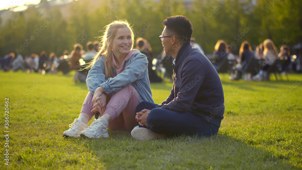 Fototapeta premium Young multiethnic couple talking and laughing sitting on grass in park
