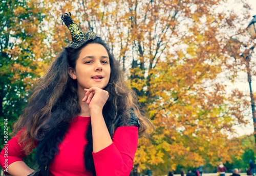 Girl brunette in a fairytale image in an autumn park. A girl in a red dress and a black vest against a yellow leaves licks her lips threatening to bite. Insidious cunning witch looking to the camera
