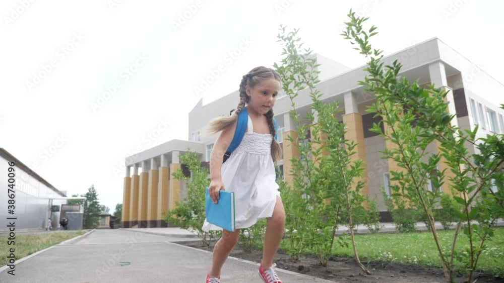 little girl kid with a backpack and a textbook runs hurrying to school. education concept. little schoolgirl with a backpack runs to the school building. child running with fun textbook back view little girl kid with a backpack and a textbook runs hurrying to school. education concept. little schoolgirl with a backpack runs to the school building. child running with fun textbook back view