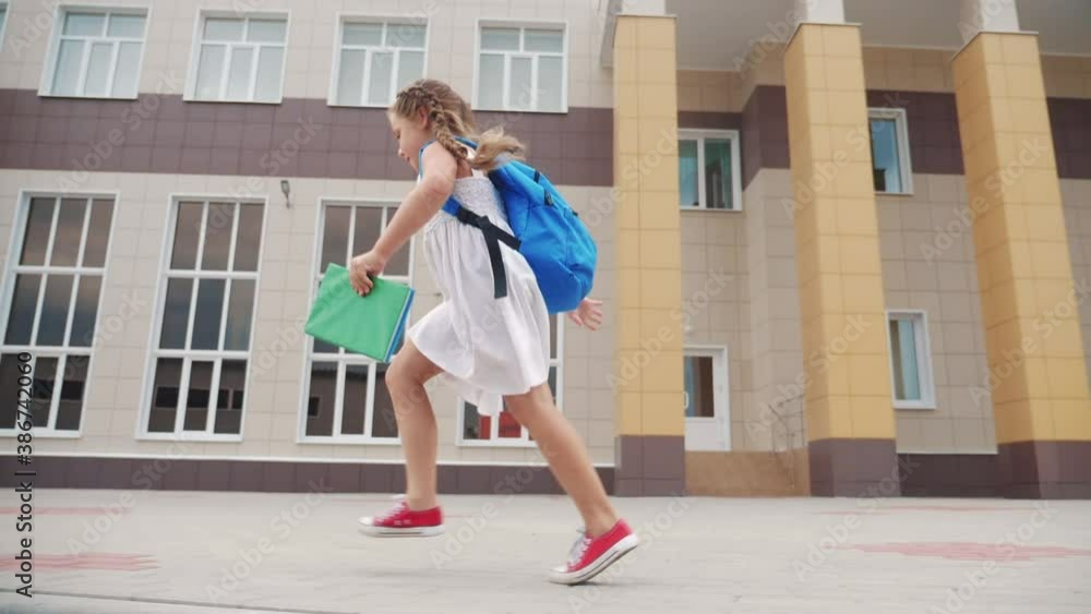 little girl kid with a backpack and a textbook runs hurrying to school. education concept fun. little schoolgirl with a backpack runs to the school building. child running with textbook back view