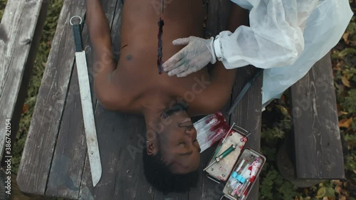 Man in coverall doing autopsy of dead afro male lying on wooden table outdoors photo