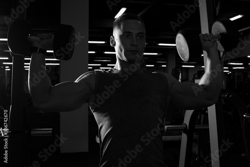 A muscular man is doing dumbbells overhead press in a gym. A bodybuilder is doing a workout with dumbbells for getting ready for the bodybuilding competition. Black and white silhouette photo.