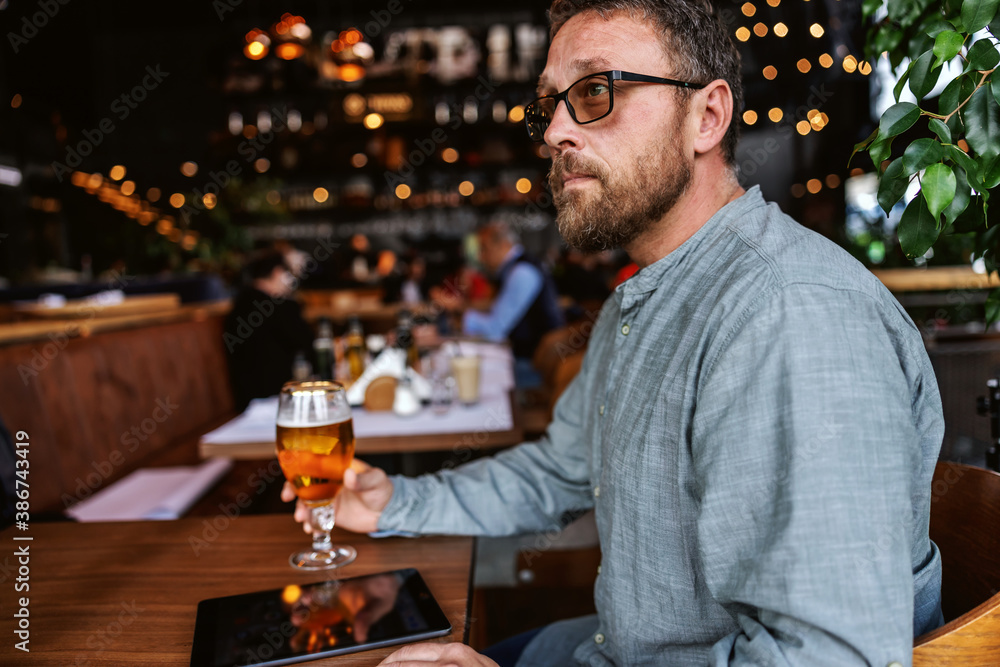 Middle aged bearded man with eyeglasses sitting in a bar, having a glass of fresh cold light beer after work.