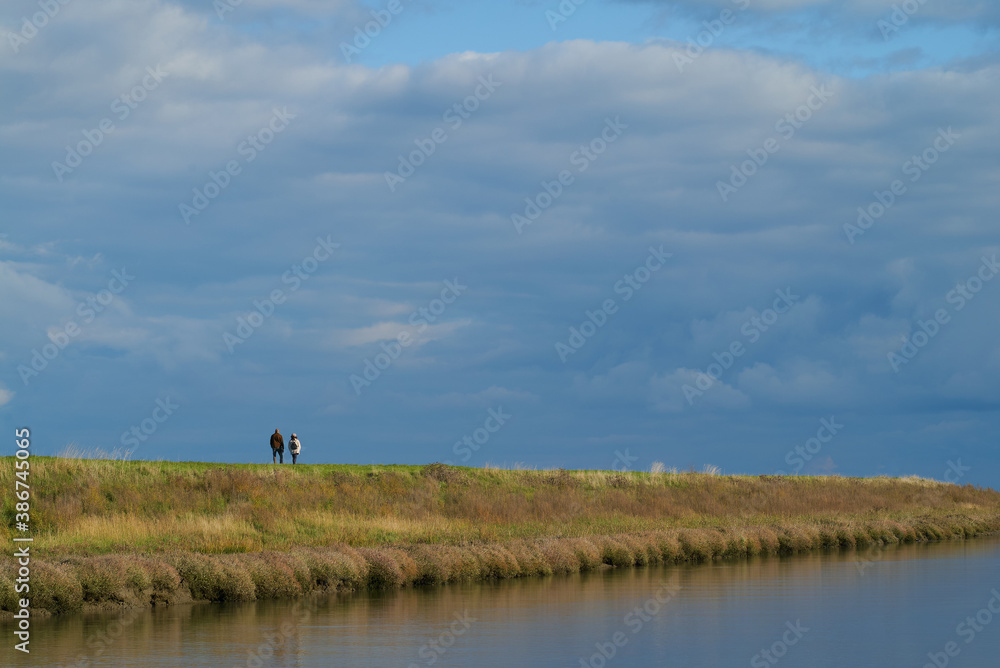 An unidentifiable couple walks on the dike at Langwarder Groden (Germany) next to a tidal way, the sky is cloudy but the sun is shining