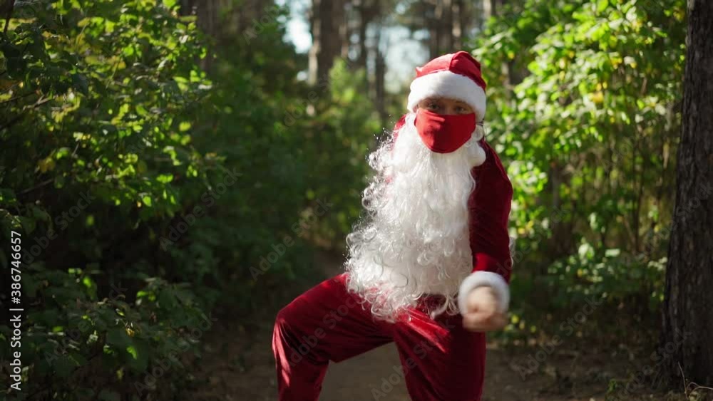 Playful man dressed in Santa Claus suit with medical mask on his face funny dancing in the forest. Christmas and coronavirus Covid-19 Quarantine during the holidays. Slow motion Playful man dressed in Santa Claus suit with medical mask on his face funny dancing in the forest. Christmas and coronavirus Covid-19 Quarantine during the holidays. Slow motion