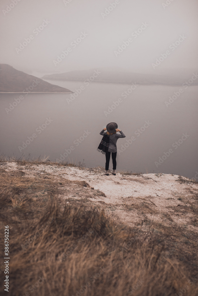 Girl traveler in a hat stands on a mountainside looks into the distance and holds a hat. the wind is blowing a scarf. Wide and smooth river. two islands away. gloomy and cold windy foggy weather