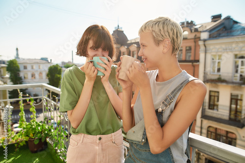 Canvas Print Two cheerful women drinking coffee or tea while standing on the balcony