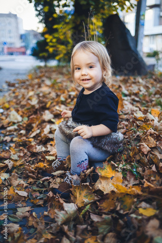 A little girl of 1.5 years is standing and smiling. The child throws leaves. Cheerful girl in the autumn forest