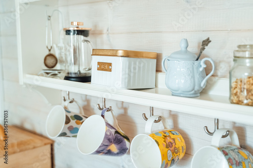 Kitchen wooden shelf with tea leaves in gold box and accessories, blue sugar bowl with spoon, strainer, press. Many colorful cups, mugs are hanging from hooks. Cozy interior in a country house