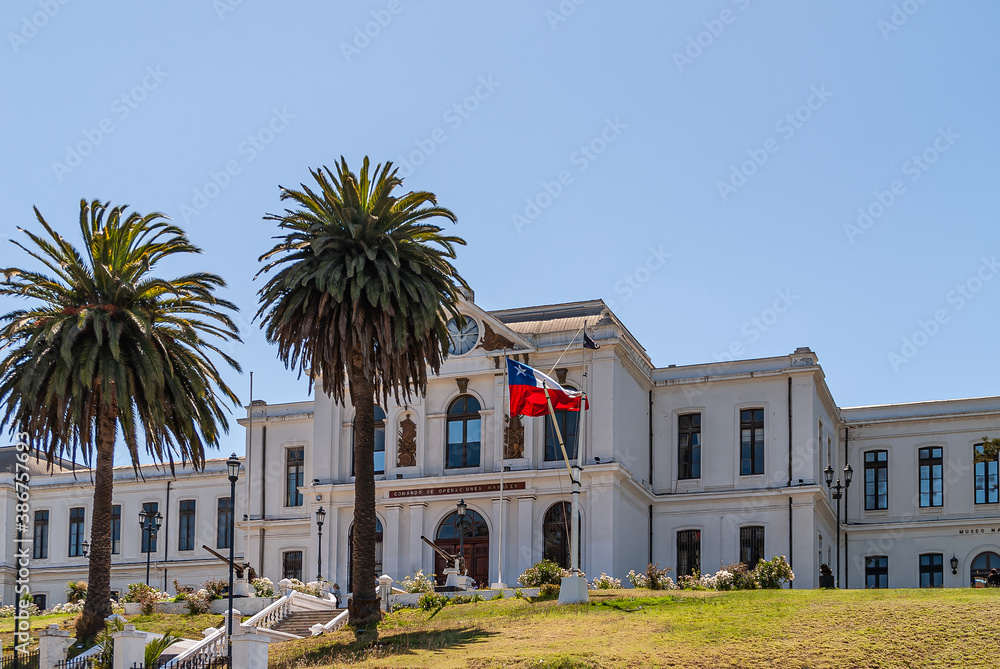 Obraz premium Valparaiso, Chile - December 8, 2008: White historic building of Navy Headquarters on green lawn hill under blue sky with couple of palm trees and a few machine guns. Flag adds color.