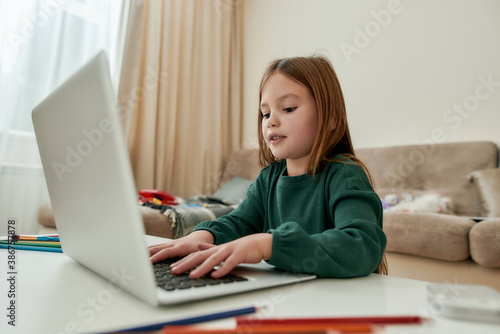 A cute small girl enjoys chatting with her friends via a laptop while sitting alone in a huge bright guestroom alone during distance education