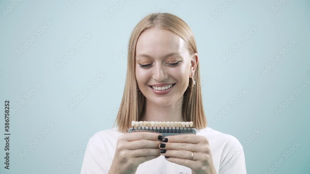 Smiling female with natural white teeth hold teeth color palette. Happy woman face with perfect smile laughing to camera in light blue background, selective focus. Smile healthy teeth concept, 4k Smiling female with natural white teeth hold teeth color palette. Happy woman face with perfect smile laughing to camera in light blue background, selective focus. Smile healthy teeth concept, 4k