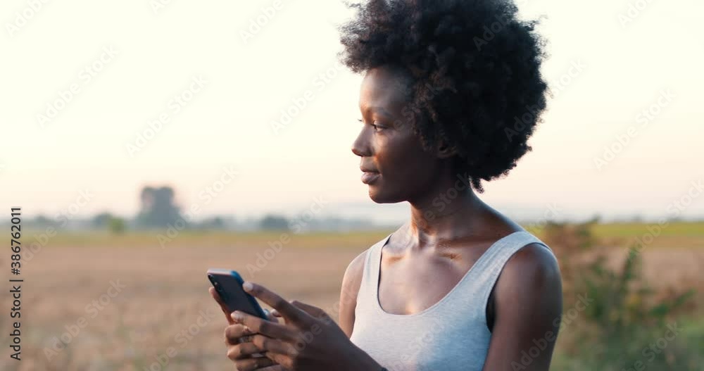 Sporty young African American woman with curly hair standing in road outdoors and tapping or scrolling on smartphone. Female texting message on mobil phone. Chatting and messaging. Cellphone. Online.