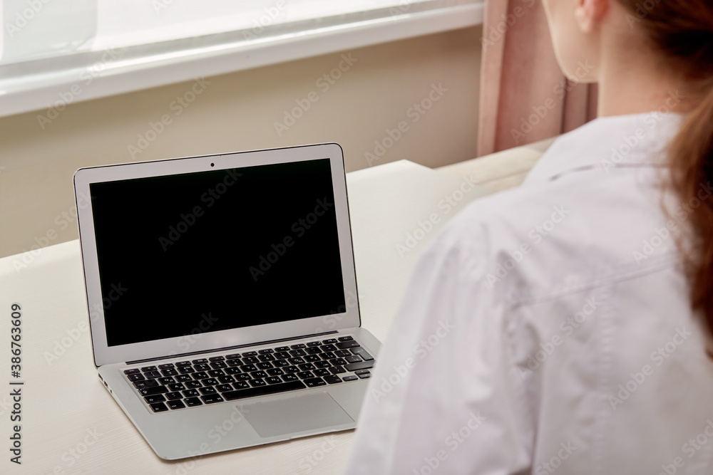 Back view of a young female doctor and a laptop with a black blank screen. Online doctor's consultation. Back view of a young female doctor and a laptop with a black blank screen. Online doctor's consultation.