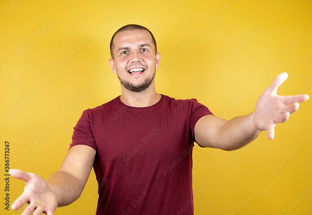Russian man wearing basic red t-shirt over yellow insolated background looking at the camera smiling with open arms for hug. Russian man wearing basic red t-shirt over yellow insolated background looking at the camera smiling with open arms for hug.