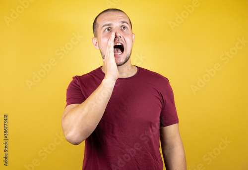 Russian man wearing basic red t-shirt over yellow insolated background shouting and screaming loud to side with hand on mouth. Communication concept.