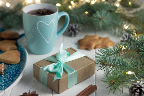 Gift with mint ribbon, cup of tea, gingerbread, next to branches of Christmas tree with light bulbs on white background