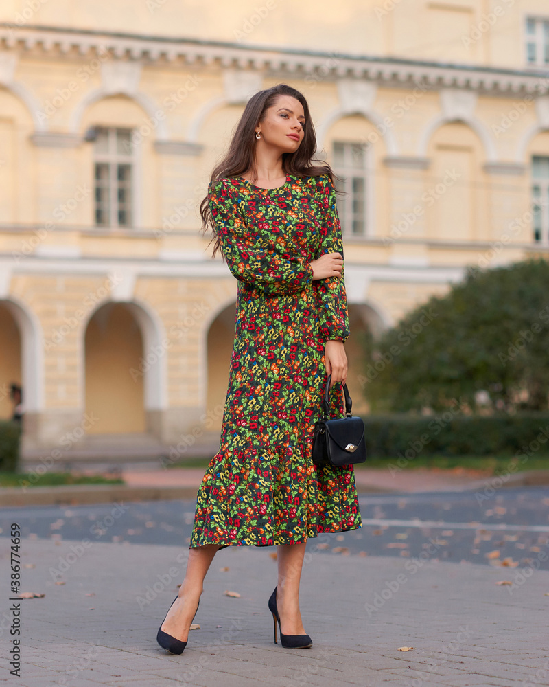Elegant woman in green dress walking city street on fall or autumn day. Pretty girl with long wavy hair holding handbag