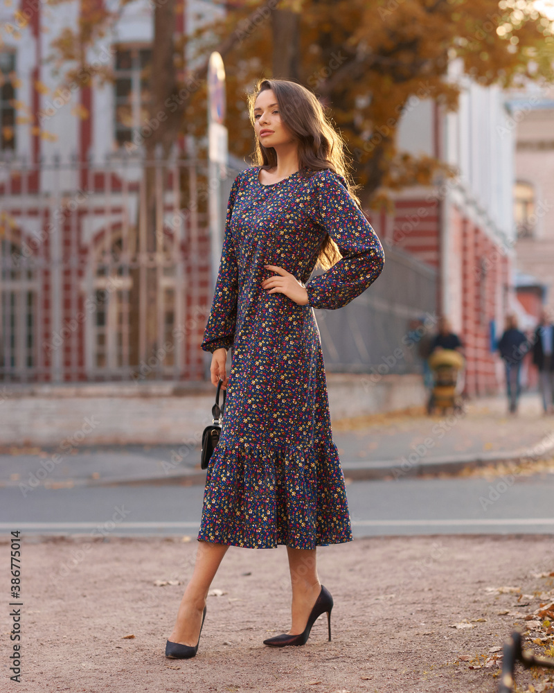 Elegant woman in purple dress walking city street on fall or autumn day. Pretty girl with long wavy hair holding handbag