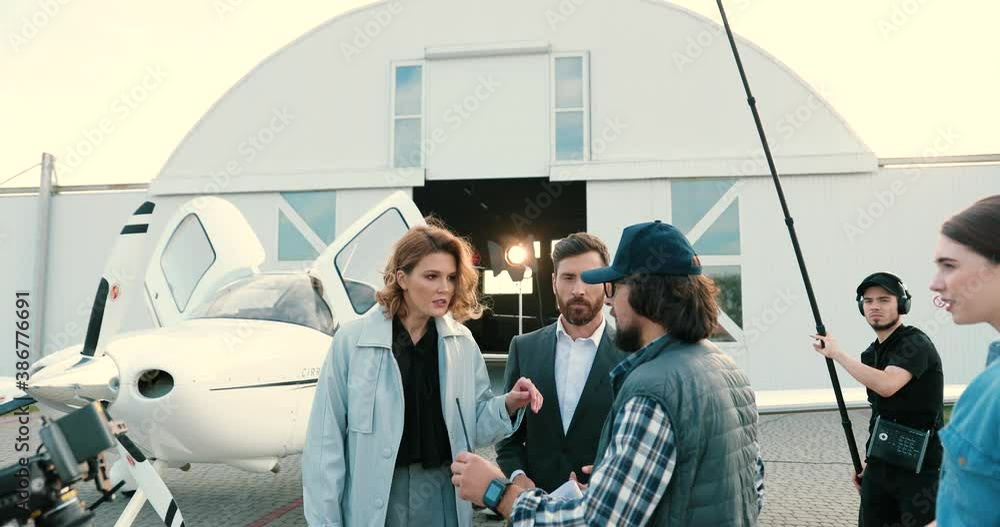 Caucasian crew on backstage of shooting outdoors. Male film director with papers talking and explaining to actors on set. Filming crew making video near hangar. Cinematography concept