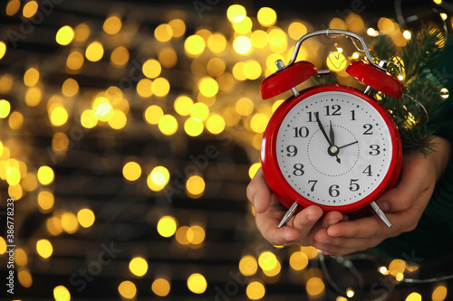 Woman holding alarm clock against blurred lights, closeup with space for text. New Year countdown