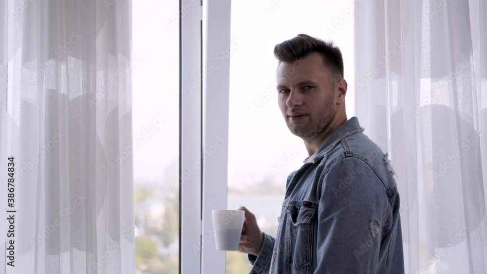 A man looks out the window and drinks delicious and fragrant coffee from a white Cup. Coffee break pause.
