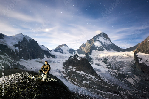 Amazing scenery in evening, man sitting on stone, beautiful mountains with white snow on background. Gorgeous mountain ridge with high rocky peaks, milky way with shining stars in sky, wonderland. © anatoliy_gleb
