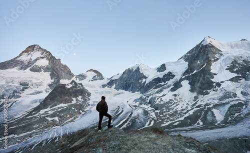 Back view of alone tourist looking at beautiful mountains scenery, moon in evening sky. Trekking, mountain hiking, man reaching peak. Wild nature with amazing views. Sport tourism in Alps.