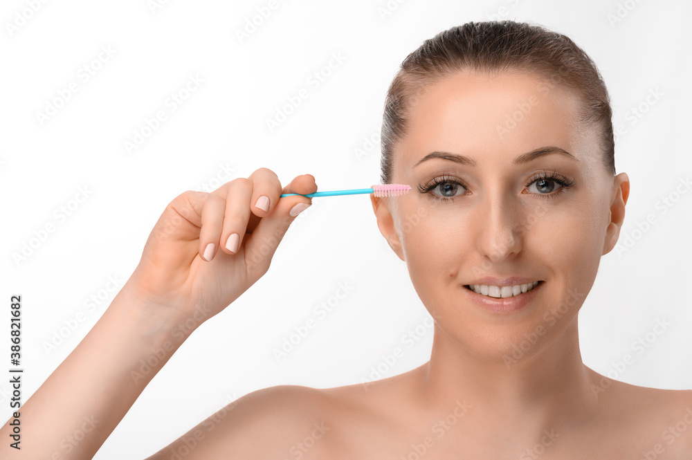 A beautiful light-skinned girl with beautiful eyes takes care of her extended eyelashes with an eyelash brush.  Soft light isolated white background.
