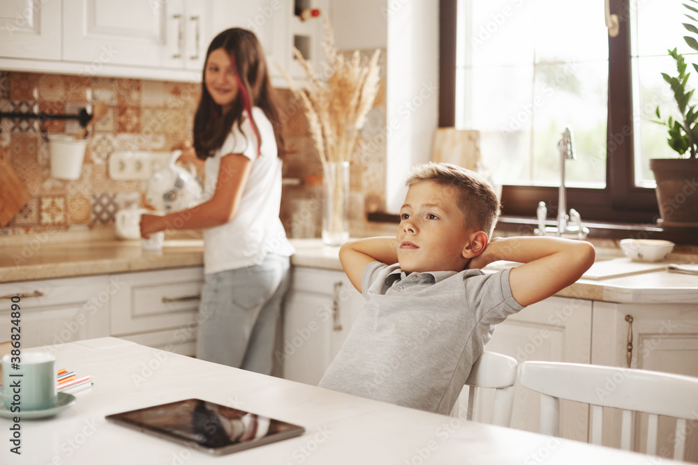 A tired boy pulls himself up after online lessons in the kitchen at home, his sister helps him. Distance learning, online education, home schooling concept A tired boy pulls himself up after online lessons in the kitchen at home, his sister helps him. Distance learning, online education, home schooling concept