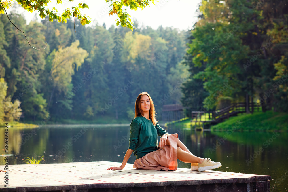 Side view of a dreaming young woman sitting on the pier of the forest and river in the background. Side view of a dreaming young woman sitting on the pier of the forest and river in the background.
