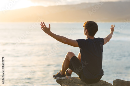 Uplifted young man feeling good sitting on top of a cliff above the ocean view and raising arms up to the sky. Hope and gratitude.