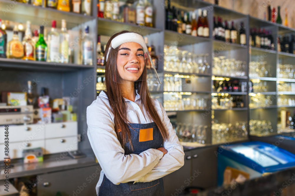 Portrait of a beautiful bartender standing at the counter smiling and looking at the camera while while wearing face shield due to covid-19, shelves full of bottles with alcohol on the background