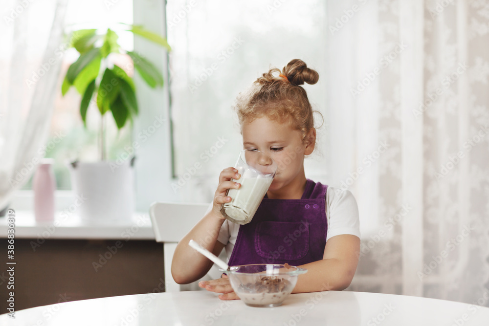 Cute little girl eats cereal with milk for Breakfast. The girl drinks milk from a glass glass. Healthy Breakfast, taking care of children. Space for text