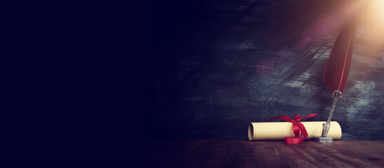 Old feather quill ink pen with inkwell and paper scroll over wooden desk in front of black wall background. Conceptual photo on history, fantasy, education and literature topic.