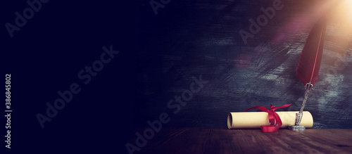 Old feather quill ink pen with inkwell and paper scroll over wooden desk in front of black wall background. Conceptual photo on history, fantasy, education and literature topic. photo