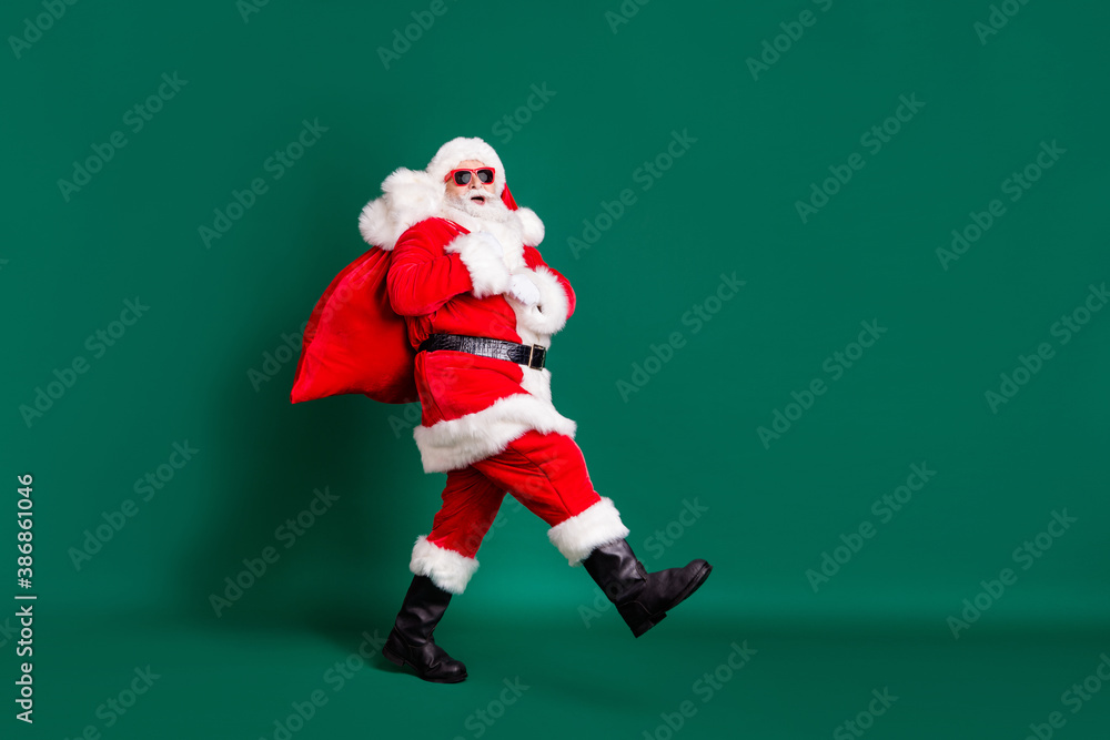 Full length profile photo of retired grandpa white beard walking bearing sack gifts children wear red santa x-mas costume coat gloves sunglass headwear isolated green color background Full length profile photo of retired grandpa white beard walking bearing sack gifts children wear red santa x-mas costume coat gloves sunglass headwear isolated green color background